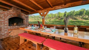 A rustic wooden dining area with a brick fireplace, a long table set for dining, and a view of a vineyard through large windows.