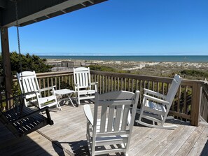 A wooden deck with white chairs and a table, overlooking a beach and ocean.