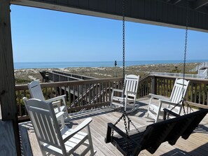 A wooden deck with white chairs and a swing, overlooking a beach and ocean.