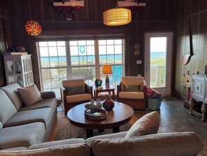 A cozy living room with a view of the beach, a round wooden table, two armchairs, and a cabinet.