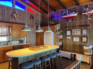 Une cuisine avec des meubles en bois, un îlot central doté de tabourets, un éclairage suspendu et des éléments de décoration sur le thème de la marine.