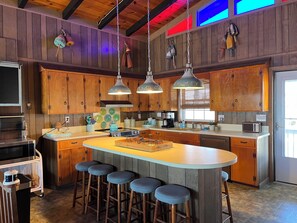 Une cuisine avec des meubles de rangement en bois, un îlot central doté de tabourets, un éclairage suspendu et un four à micro-ondes.