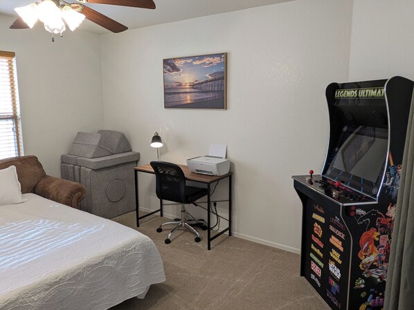 Un dormitorio con cama, escritorio con impresora, ventilador de techo, un cuadro en la pared y una consola de videojuegos.