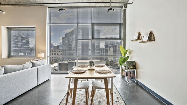 A modern living room with a dining table, sofa, and large windows offering a city view. - *NEW* Trendy & Central Gulch Condo! (Nashville)