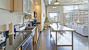 A modern kitchen with stainless steel appliances, a dining area with a long table, and a view of a cityscape through large windows. - Stylish 2 Bedroom in the Gulch (Nashville)