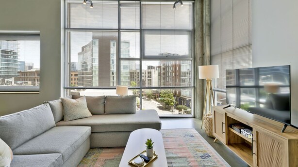 A modern living room with a grey sofa, a coffee table with a plant, a TV on a wooden stand, and large windows with city views. - Stylish 2 Bedroom in the Gulch (Nashville)