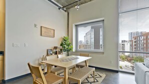 A modern dining area with a wooden table, chairs, and a bench. There is a large window with blinds, a potted plant, and a view of buildings outside. - Stylish 2 Bedroom in the Gulch (Nashville)