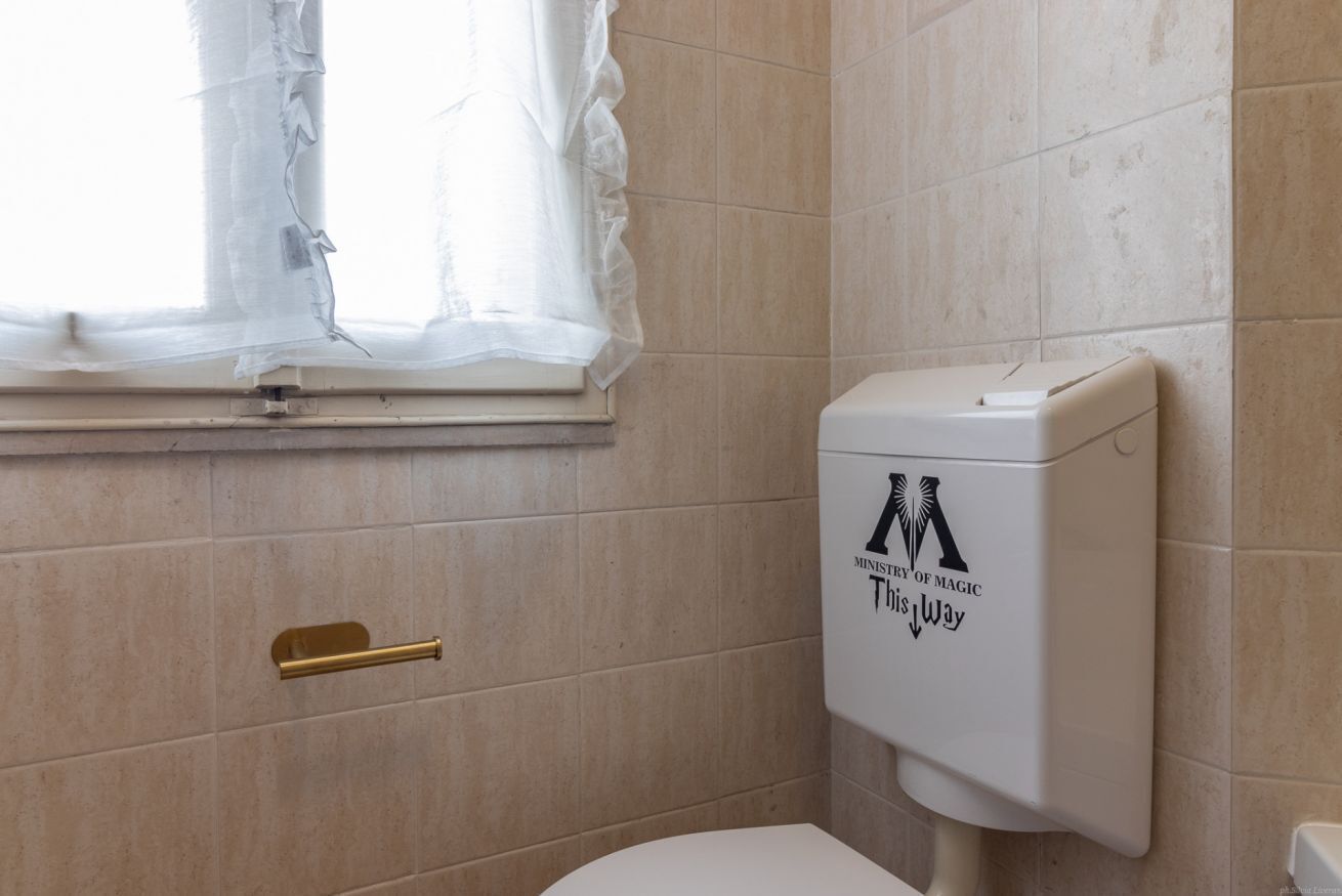 A bathroom with beige tiles, a white toilet, a gold handle, and a window with a white curtain.