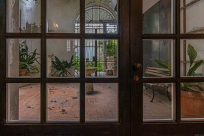 Une vue à travers une fenêtre à carreaux, avec des plantes en pot, un banc et un chemin pavé de briques.