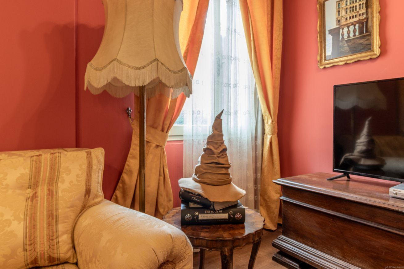 Una habitación con sofá, lámpara, libros apilados, televisor y ventana con cortinas.