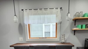 Ein Essbereich mit einem Holztisch und zwei Stühlen, einem Fenster mit Jalousien, einem Regal mit Gläsern und einem kleinen schwarzen Gerät.