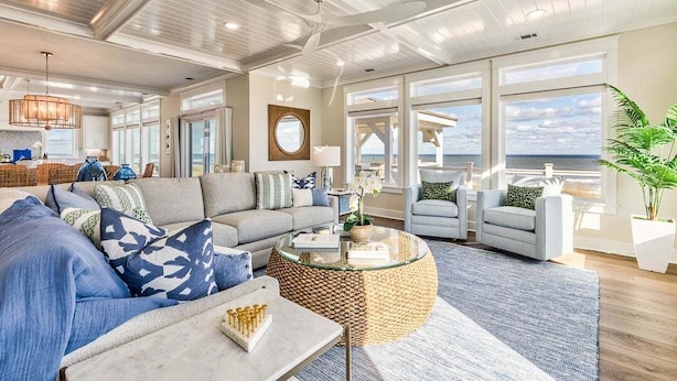A spacious living area with a large sofa, a round coffee table, and a view of the ocean through large windows. - Pura Vida | Oceanfront | Private Pool, Hot Tub, and Dog Friendly (Virginia Beach)