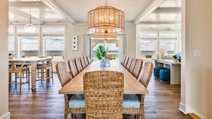 A spacious dining area with a long wooden table and wicker chairs, a hanging light fixture, and large windows offering a view of the outdoors. - Pura Vida | Oceanfront | Private Pool, Hot Tub, and Dog Friendly (Virginia Beach)