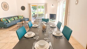 Un'area pranzo con tavolo apparecchiato per sei persone, circondata da sedie blu, un divano con cuscini colorati e una TV sullo sfondo.