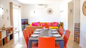 A dining area with a long table set for six, surrounded by orange chairs. A sofa with colorful cushions is against the wall, and a television is visible in the background.