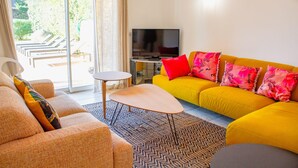 A living room with a yellow sofa, orange armchair, and a small round table. A TV is mounted on the wall, and there is a sliding glass door leading to an outdoor area with sun loungers.