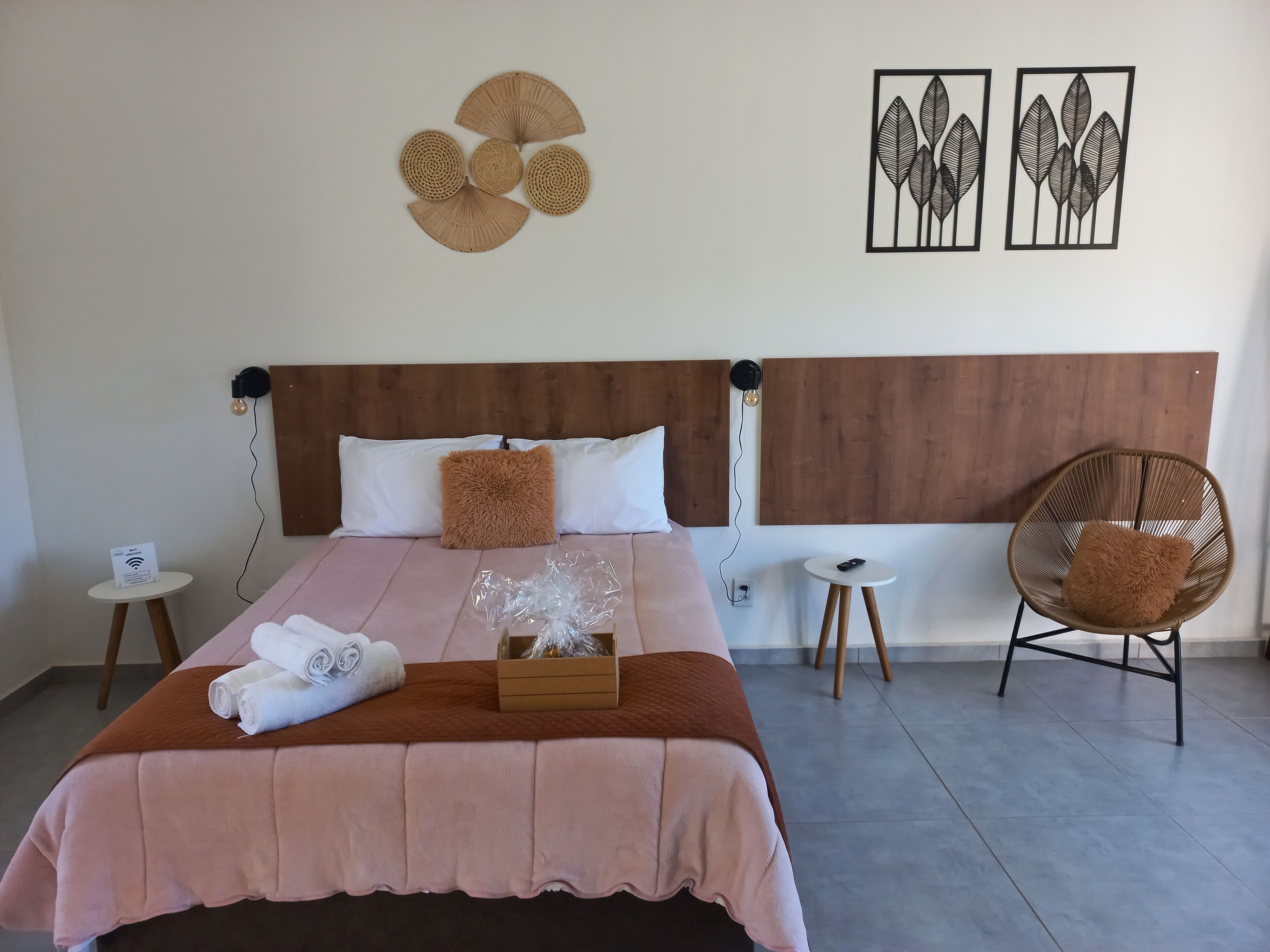 A neatly made bed with a brown headboard, a wooden side table with a lamp, a wicker chair, and artwork on the wall.