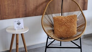A wicker chair with a fluffy pillow against a wooden wall, a small table with a Wi-Fi sign, and a tiled floor. - EMBAHÚ SUÍTES  (Santo Antonio do Pinhal)