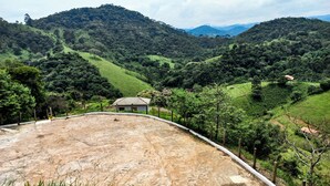 A rural landscape with a small house surrounded by lush green hills and a dirt path. - EMBAHÚ SUÍTES  (Santo Antonio do Pinhal)