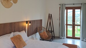 A hotel room with a large bed, a wooden headboard, a wicker chair, a ladder, and a view of greenery through the door. - EMBAHÚ SUÍTES  (Santo Antonio do Pinhal)