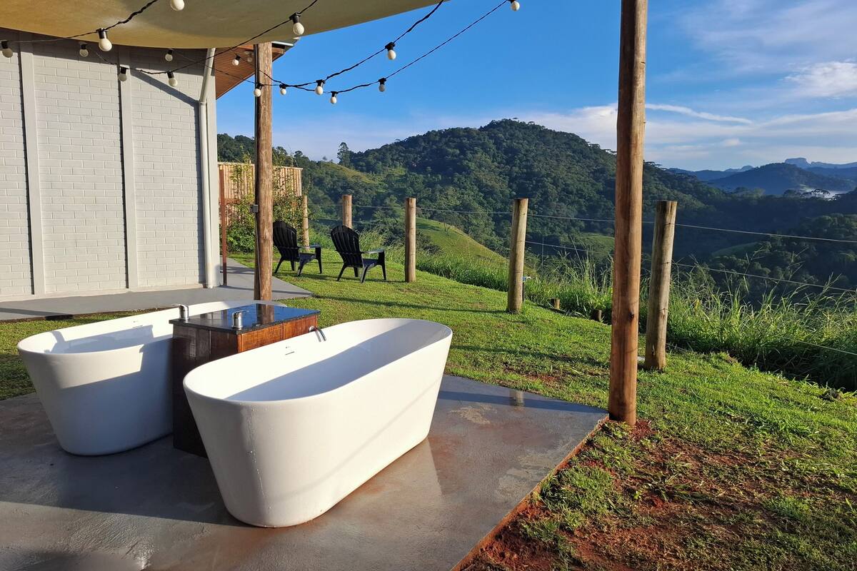 A modern outdoor bathroom with two tubs, a wooden table, and chairs, set against a backdrop of lush greenery and hills.