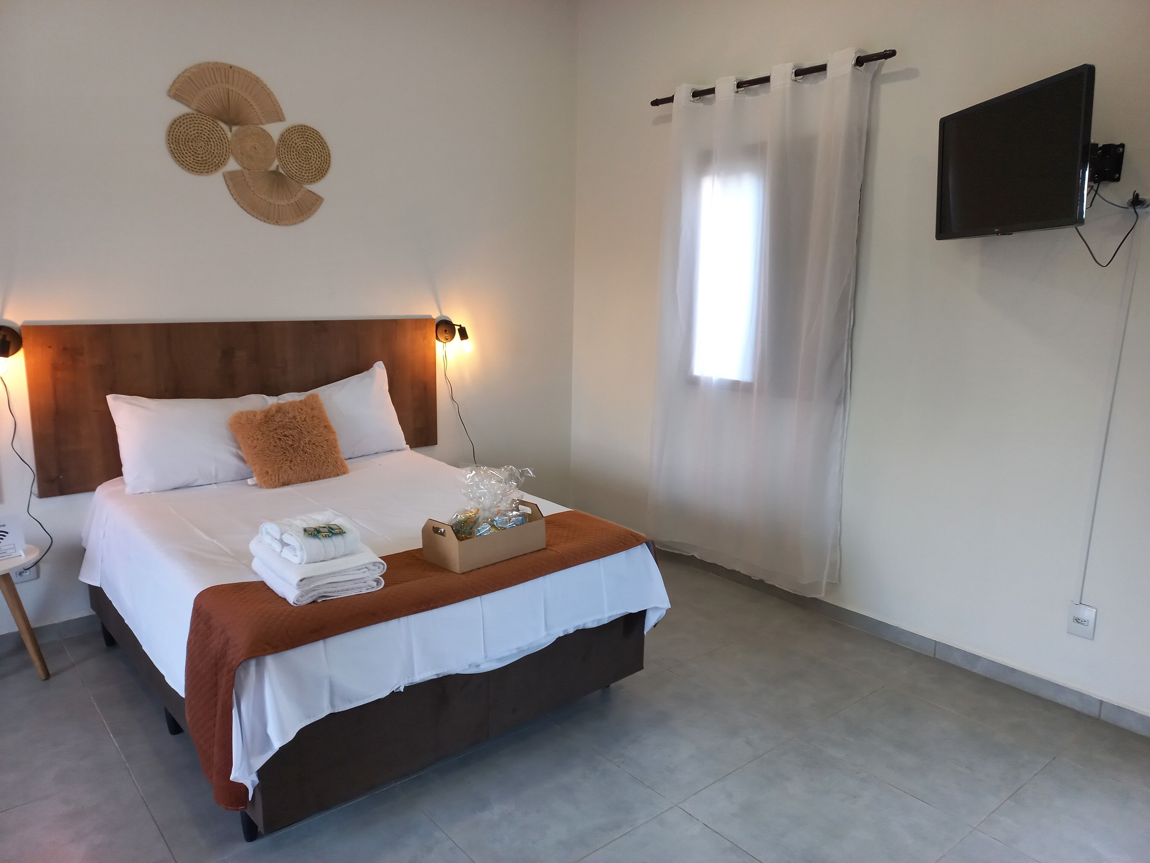 A neatly made bed with a brown headboard, white linens, and a brown throw blanket. There is a wooden nightstand with a lamp and a small table with a towel and a box of toiletries. A flat-screen TV is mounted on the wall, and a window with sheer curtains is visible.