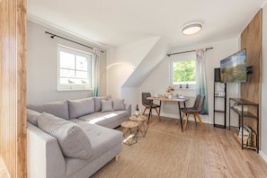 En moderne stue med en grå sofa, et spisebord med stole, et TV ophængt på væggen og et vindue med gardiner.
