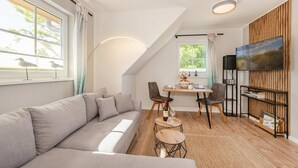 A modern living room with a grey sofa, a dining table with chairs, a TV on the wall, and a bookshelf.