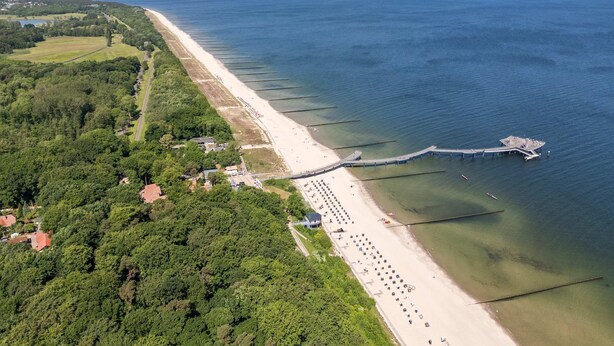 An aerial view of a coastal area with a beach, a pier, and surrounding greenery.