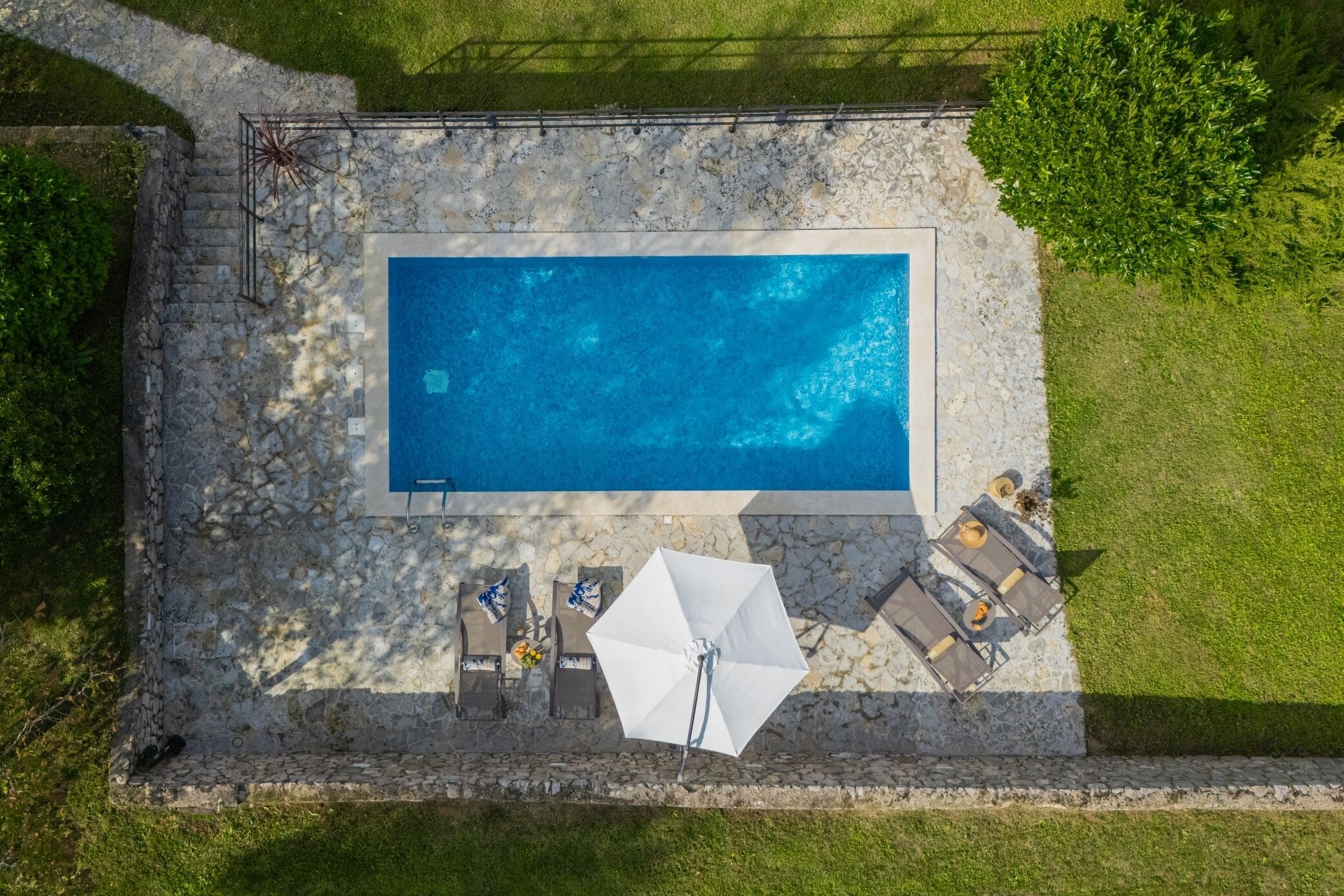 Vista aérea de una piscina rectangular con borde de piedra, rodeada por un solárium de piedra y equipada con sillas de descanso y sombrillas.