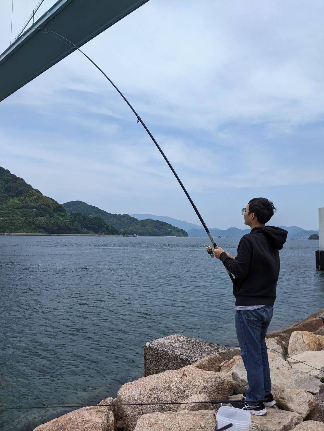 A person fishing by a rocky waterfront with a view of distant hills.