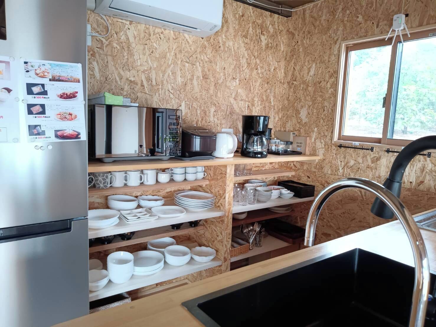 Cocina compacta con refrigerador de acero inoxidable, microondas, cafetera y varios platos y utensilios en estantes.