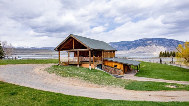 A wooden cabin with a porch and a stone foundation, overlooking a lake and mountains.