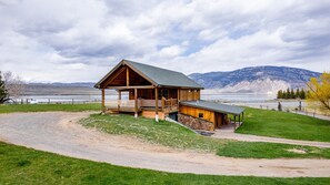 A wooden cabin with a porch and a stone foundation, overlooking a lake and mountains.