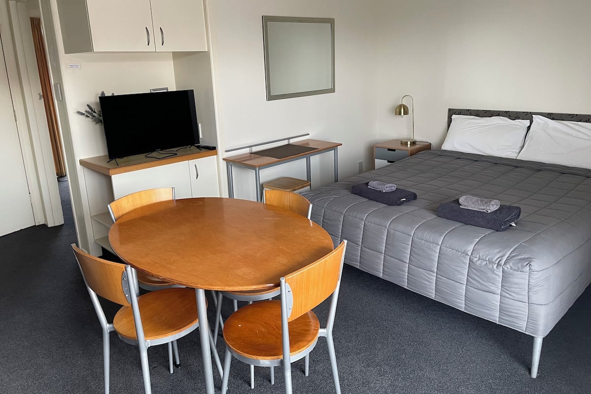 A hotel room with a bed, a desk with a TV, a chair, a table, and a mirror.