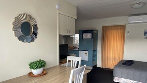 Ein gemütlicher Raum mit Esstisch aus Holz, weißen Stühlen, einer kleinen Topfpflanze, einem Bett mit floralem Überwurf, einem blauen Schrank und einem Fernseher.