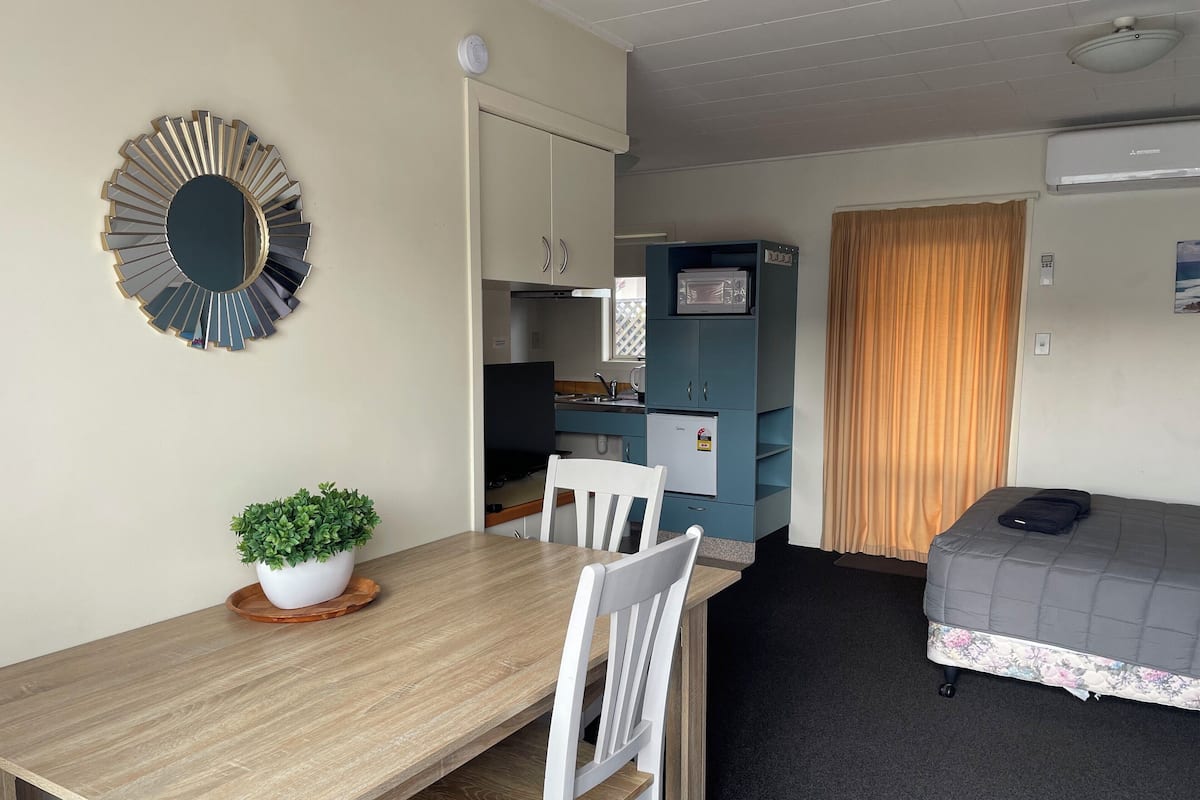 A compact room with a wooden dining table, white chairs, a small potted plant, a bed with a floral bedspread, a blue cabinet, and a television.