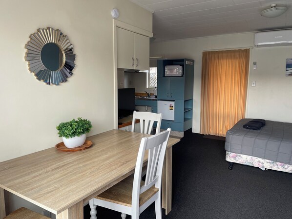 A compact room with a wooden dining table, white chairs, a small potted plant, a bed with a floral bedspread, a blue cabinet, and a television. - Bella Tuscany On Kennedy (Napier)