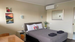 Ein Hotelzimmer mit einem Bett, einem Schreibtisch, einem Sessel, einem Nachttisch, einer Vase mit Pflanze und zwei Wandklimageräten.