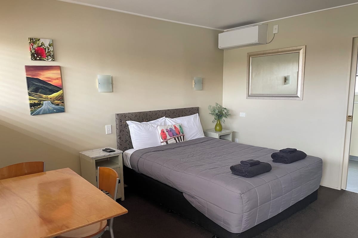 A hotel room with a bed, a desk, a chair, a nightstand, a vase with a plant, and two wall-mounted air conditioning units.