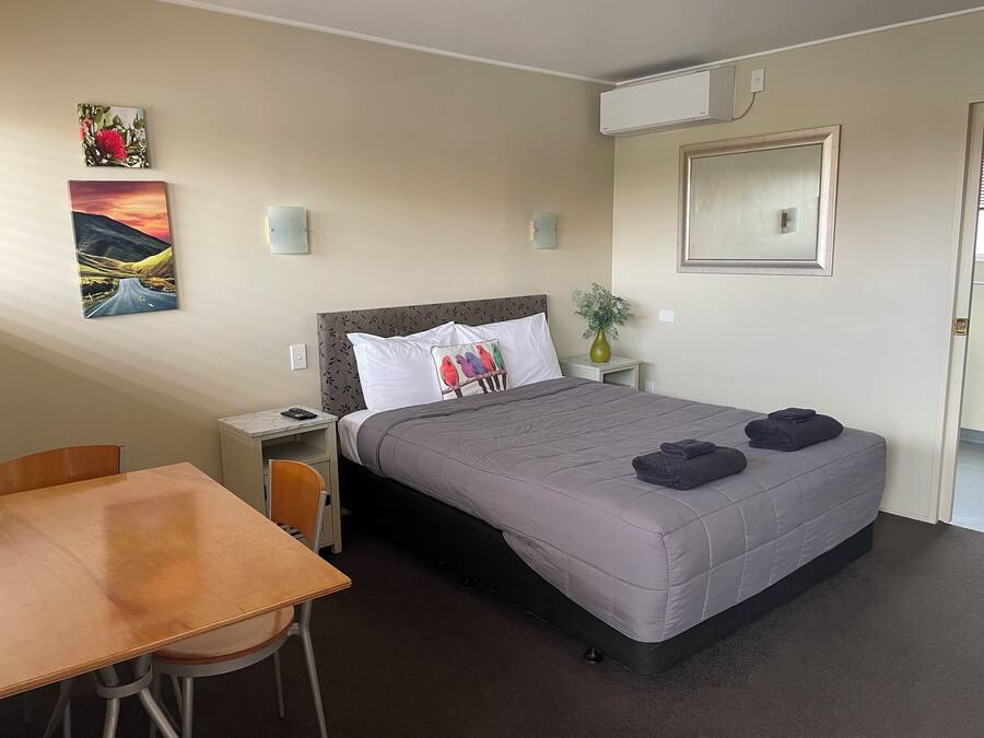 A hotel room with a bed, a desk, a chair, a nightstand, a vase with a plant, and two wall-mounted air conditioning units.