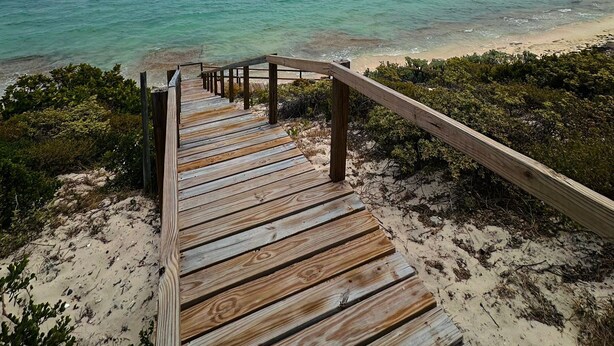 Un escalier en bois mène à une plage bordée d’une eau turquoise et limpide, sous un ciel nuageux.