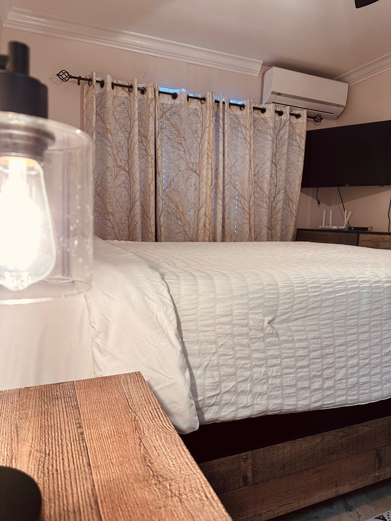 Ein Schlafzimmer mit einem Bett, einem Nachttisch aus Holz, einer Lampe, einer Wand-Klimaanlage und einem Fernseher.