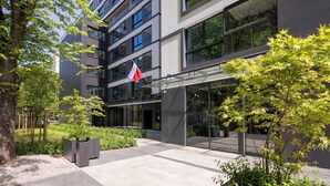 Een modern gebouw met een glazen ingang, omringd door groen en een vlag.