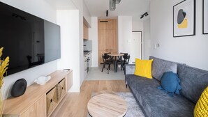 Een moderne woonkamer met een flatscreen televisie, een houten dressoir, een grijze bank met gele en blauwe kussens, en een eethoek met een tafel en stoelen.
