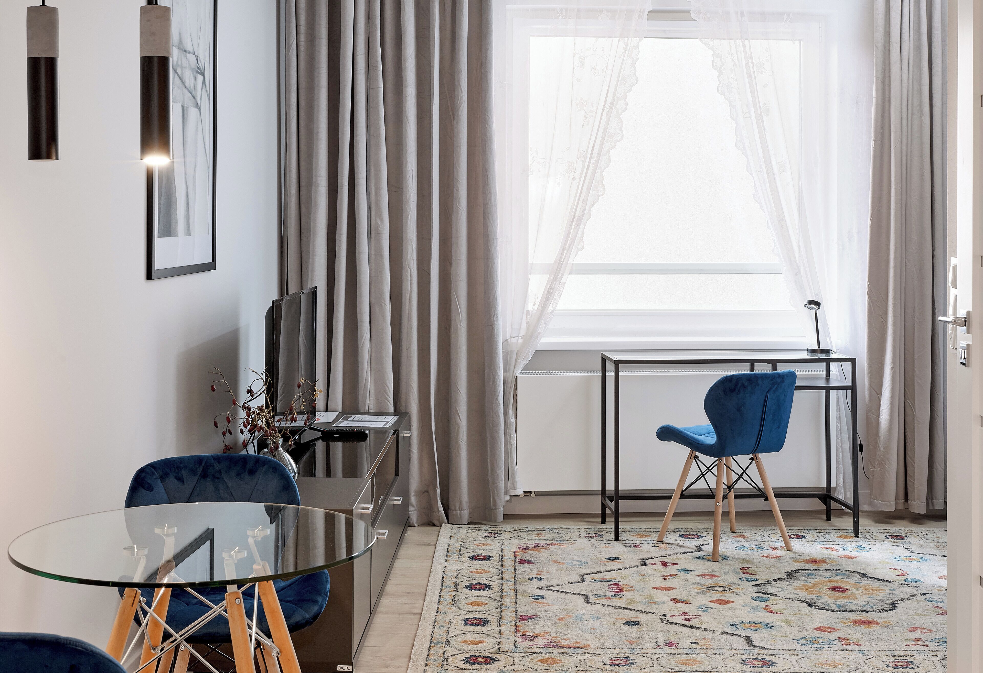 Ein modernes Wohnzimmer mit Glastisch, blauen Stühlen, einem gemusterten Teppich und einem großen Fenster mit transparenten Vorhängen.