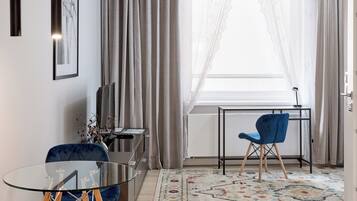 Ein modernes Wohnzimmer mit Glastisch, blauen Stühlen, einem gemusterten Teppich und einem großen Fenster mit transparenten Vorhängen.