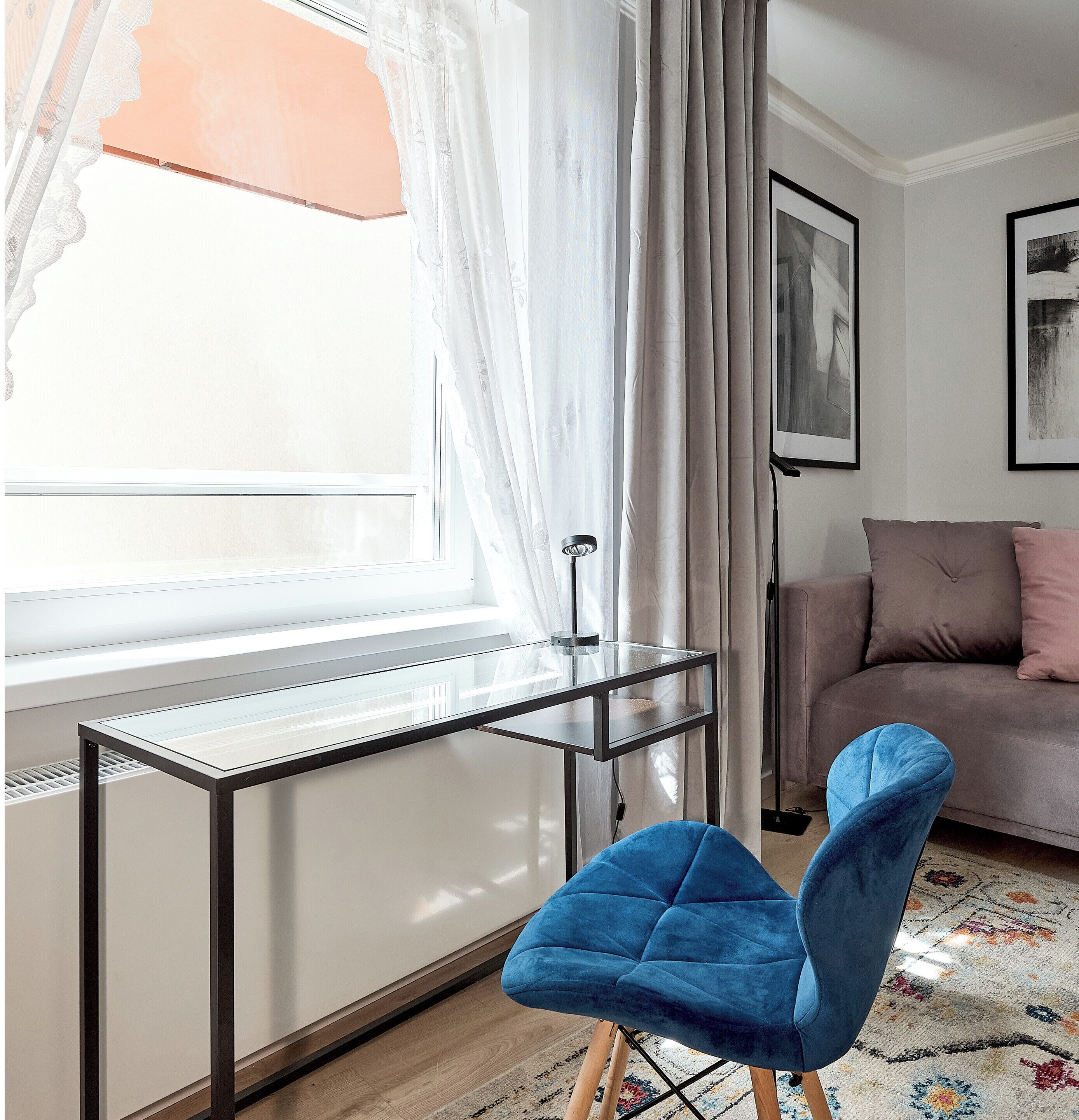 A modern living room with a glass desk, a blue chair, a sofa, and a large window with sheer curtains.