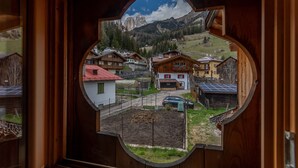 Vista attraverso una finestra con cornice in legno su un villaggio con case, un'auto e montagne sullo sfondo.