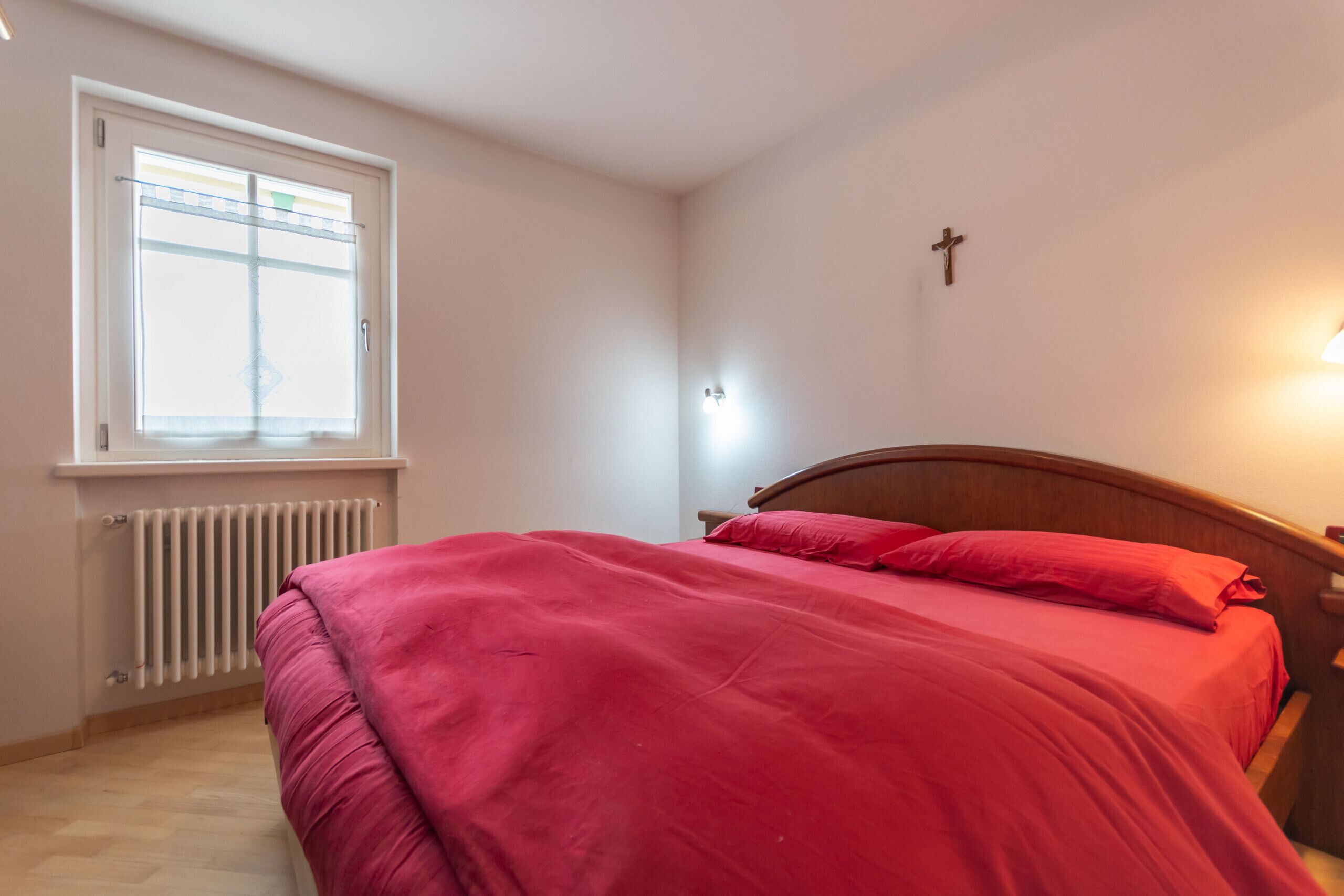 A bedroom with a bed, a window, a radiator, and a cross on the wall.
